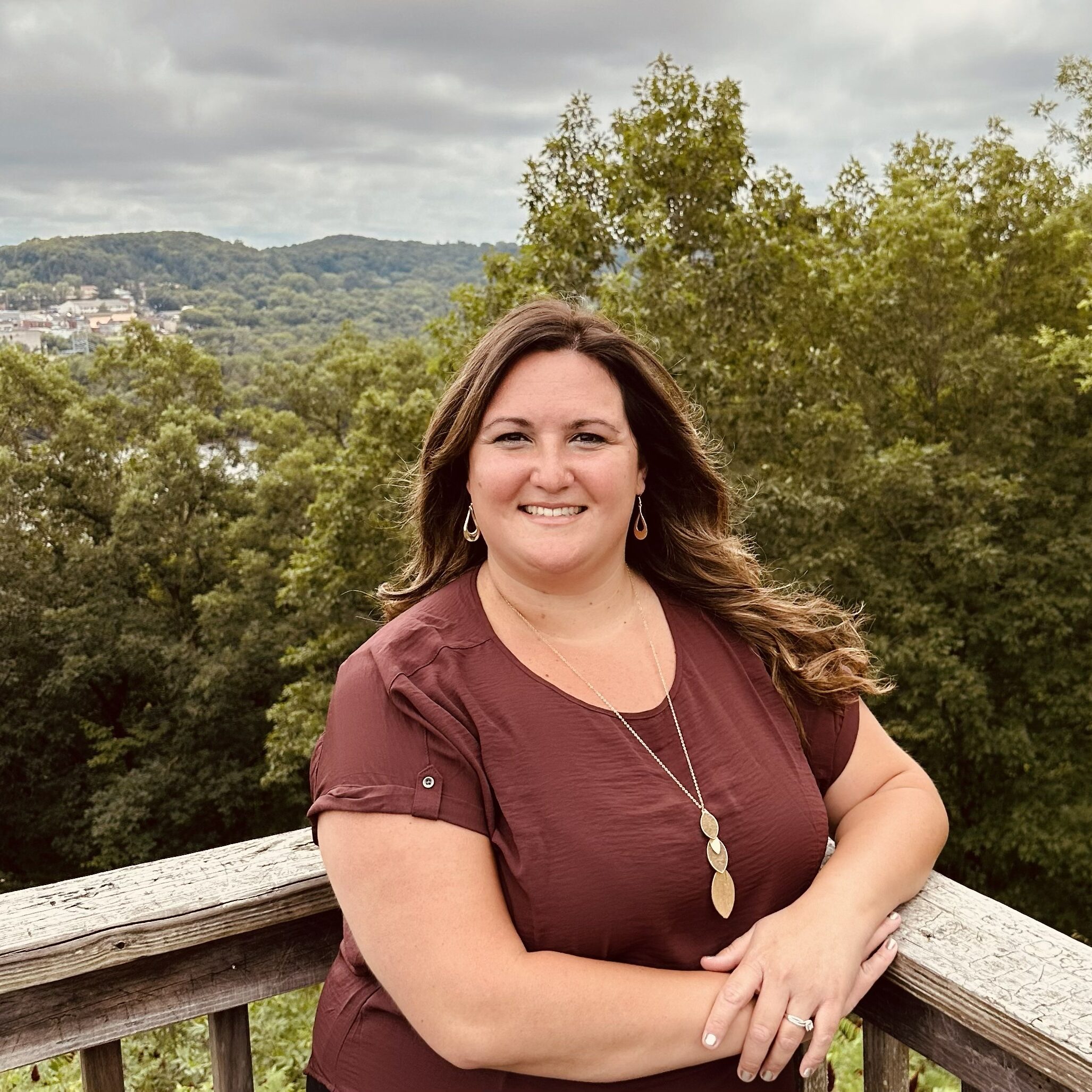Smiling woman leaning on a weathered wooden railing with trees and hillside in the background on a cloudy day
